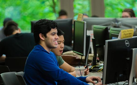 Students looking at a computer monitor and laughing with window view of forest in the background