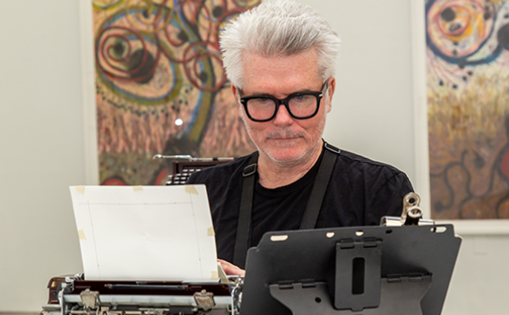 Tim Youd typing on a manual typewriter, surrounded by his paintings