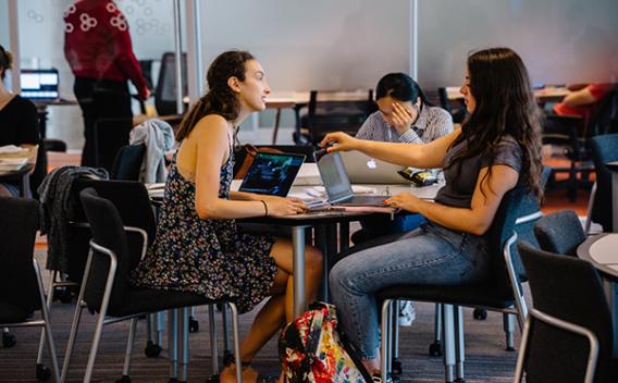 Two students sitting at table with their laptops, in discussion